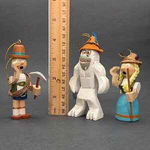 Puede incluir: Tres adornos de madera que incluyen un escalador con un piolet, un yeti blanco con un sombrero naranja y una figura con trenzas rubias, junto a una regla de madera que muestra una altura de unos 15 cm para la figura m&aacute;s alta.