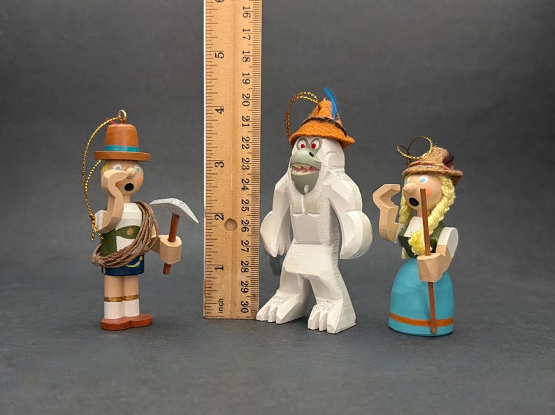 Puede incluir: Tres adornos de madera que incluyen un escalador con un piolet, un yeti blanco con un sombrero naranja y una figura con trenzas rubias, junto a una regla de madera que muestra una altura de unos 15 cm para la figura m&aacute;s alta.