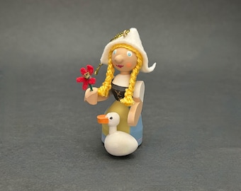 Puede incluir: Una figura de madera de una persona con gorro blanco y pelo amarillo trenzado, que sostiene una flor roja. El personaje lleva un corpiño negro sobre una falda amarilla y azul y está junto a un pequeño pato blanco con pico naranja.