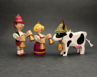 May include: Three wooden ornaments featuring a person in a red hat, a person with blonde hair, and a white cow with black spots. The two figures wear colorful clothing and hold small gold bells, while the cow wears a green hat and a bell.