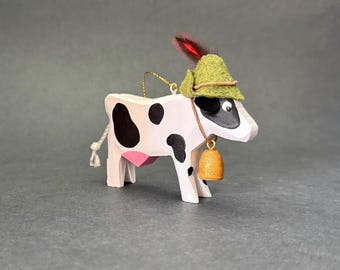 Puede incluir: Una figura de vaca de madera pintada de blanco con manchas negras, con una ubre rosa y una cola de cuerda blanca. La vaca lleva un sombrero de fieltro verde con una pluma roja y un collar de cordón marrón con un pequeño cencerro dorado.