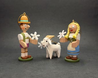 May include: Three hand-painted wooden ornaments featuring a figure in a yellow hat and green vest, a figure with blonde braided hair in a green bodice and blue skirt, and a white goat. Each figure stands on a green base with a gold hanging loop.