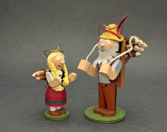 May include: Two wooden figurines of a person with blonde braided hair in a red skirt and a bearded person in a yellow hat, both carrying bundles of wooden sticks on their backs. Each figure stands on a round green base with a gold hanging loop.