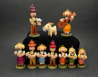Puede incluir: Una colección de diez figuras de madera pintadas a mano que representan a un grupo de personas con vestimenta tradicional, incluyendo una persona con una guitarra y una cabra. Cada figura tiene un lazo dorado, ropa colorida y una base verde.
