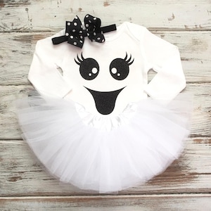 May include: A white long-sleeve bodysuit featuring a black glittery ghost face graphic with large eyes and eyelashes. The bodysuit is paired with a white tulle skirt attached at the waist and a black headband with a white polka dot bow.