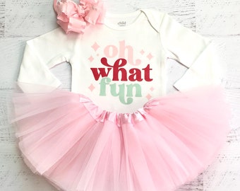 May include: A white long-sleeved onesie with the text "oh what fun" in red, green, and pink. The onesie is paired with a pink tulle tutu and a pink bow headband.
