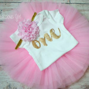K&ouml;nnte beinhalten: Ein rosa T&uuml;ll-Tutu-Rock mit einem wei&szlig;en Strampler, auf dem "eins" in goldenen Glitzer steht. Der Strampler hat eine rosa Blume auf der Schulter.