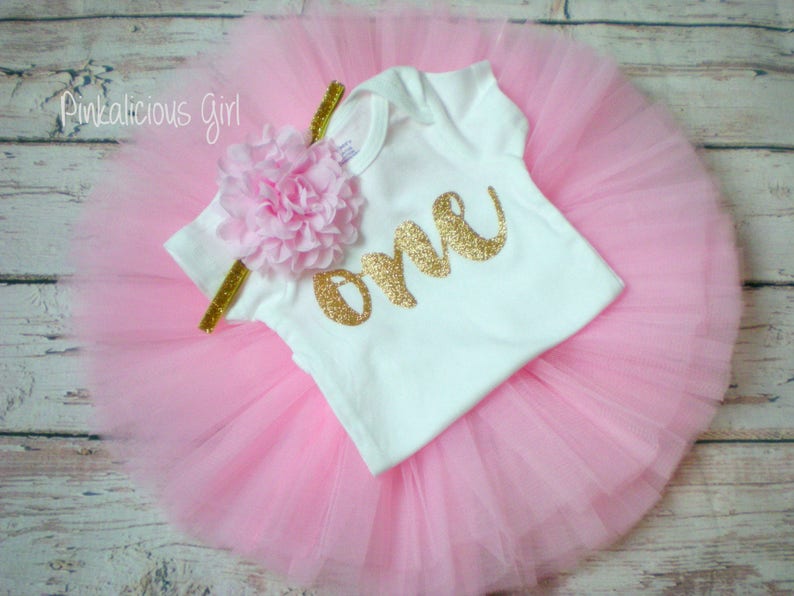 K&ouml;nnte beinhalten: Ein rosa T&uuml;ll-Tutu-Rock mit einem wei&szlig;en Strampler, auf dem "eins" in goldenen Glitzer steht. Der Strampler hat eine rosa Blume auf der Schulter.