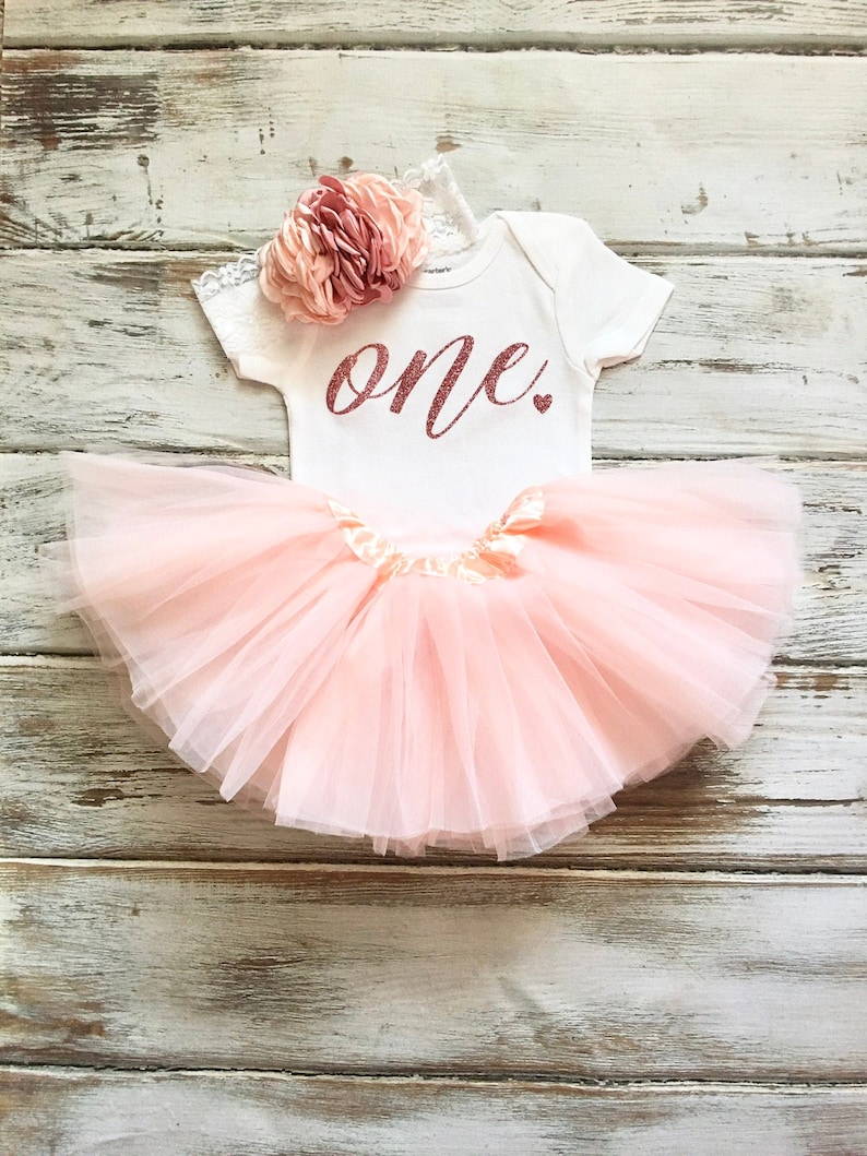 May include: A pink tulle tutu skirt with a white onesie that says "one" in rose gold glitter. The onesie has a pink flower headband.