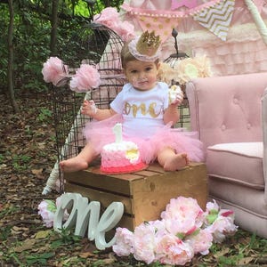 K&ouml;nnte beinhalten: Ein kleines M&auml;dchen in einem wei&szlig;en Strampler mit goldener Aufschrift "one" und einem rosa Tutu sitzt auf einer Holzkiste mit einem rosa und wei&szlig;en Kuchen vor ihr. Sie tr&auml;gt eine goldene Krone und hat Zuckerguss im Gesicht. Der Hintergrund ist mit rosa und goldenen Dekorationen geschm&uuml;ckt.