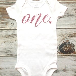 Op de afbeelding: Witte baby romper met het woord "one" in roze glitter letters en een klein roze glitter hartje.