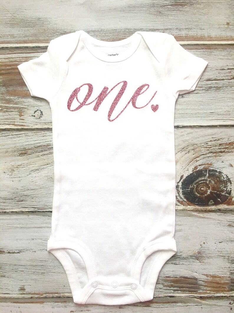 Op de afbeelding: Witte baby romper met het woord "one" in roze glitter letters en een klein roze glitter hartje.