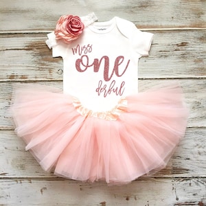 May include: A white baby bodysuit with the words "Miss One Derful" in pink glitter lettering, paired with a light pink tulle tutu skirt and a matching headband with a pink flower.