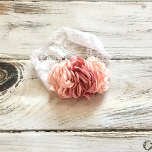 Op de afbeelding: Een roze en witte bloemenhoofdband met een witte kanten basis. De hoofdband is gemaakt van stof en heeft een gerimpeld ontwerp.