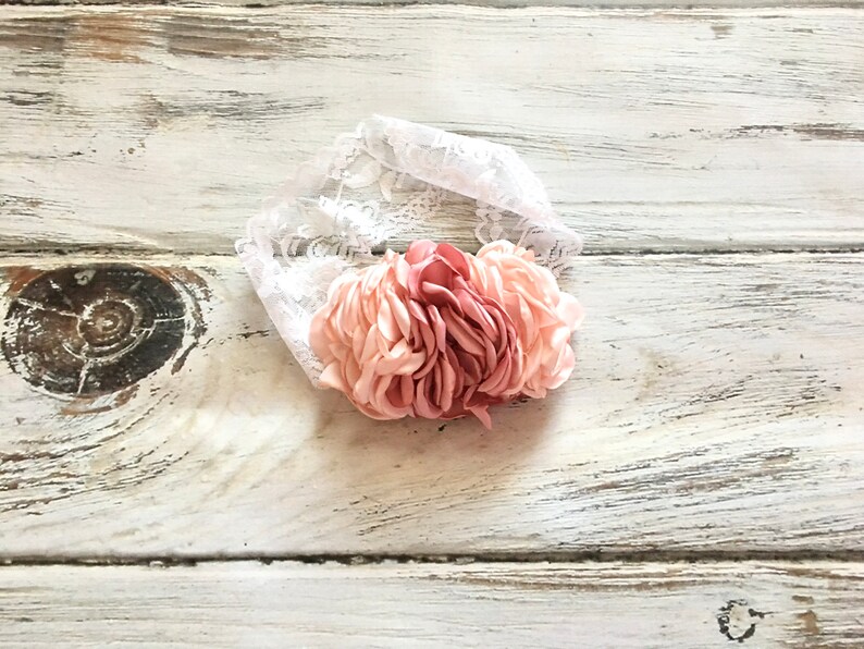 Op de afbeelding: Een roze en witte bloemenhoofdband met een witte kanten basis. De hoofdband is gemaakt van stof en heeft een gerimpeld ontwerp.