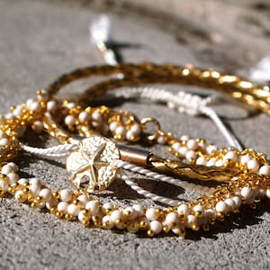 May include: Close-up of several gold and white beaded bracelets. One bracelet features a gold starfish charm. The bracelets are arranged on a textured surface, with a soft focus background.