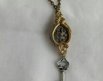 Puede incluir: Un collar de oro con un colgante de llave. La llave está unida a un medallón en forma de corazón con un mecanismo de relojería visible.