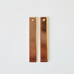 May include: Two copper-colored rectangular metal bars with a small hole at the top. The bars are positioned vertically against a white background. The bars are likely components for jewelry making or other crafts.