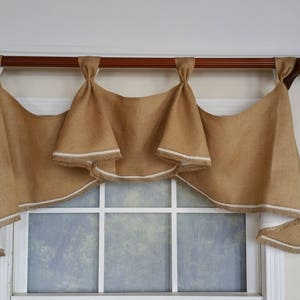 May include: A brown burlap valance with white trim hangs from a wooden rod. The valance is gathered in the center and has a scalloped edge. The valance is hung over a window with white trim.