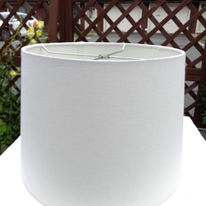 May include: A cylindrical lamp shade featuring a solid white fabric exterior with a fine, textured weave. The top opening reveals a silver-toned metal spider fitter assembly with four spokes meeting at a central ring for lamp attachment.