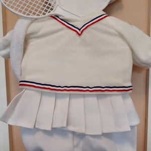 May include: A white tennis outfit with a matching visor and a tennis racket. The outfit includes a collared shirt with red and blue trim, a pleated skirt, and matching shorts. A white towel is draped over the shoulder.