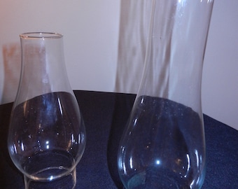 May include: Two clear glass lamp chimneys of different heights. Both chimneys feature a bulbous base that tapers into a narrower, cylindrical top section. The glass is transparent and smooth, designed to fit over a lamp burner for protection.