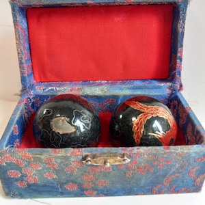 May include: A set of two black, decorative Chinese health balls with intricate designs, resting in a blue and red fabric-lined box. The box has a gold clasp and is decorated with a floral pattern.