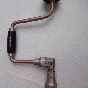 Puede incluir: Un taladro manual vintage con un mango negro y un marco de metal marrón oscuro. El taladro tiene un mango circular negro en la parte superior y un portabrocas de metal en la parte inferior. El mango dice "Companion".