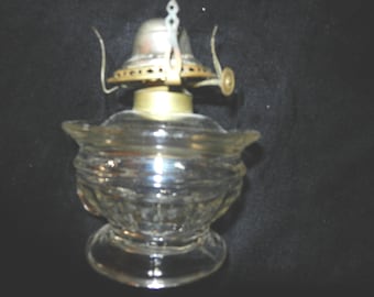 May include: A clear glass oil lamp base featuring a textured, patterned surface with a wide, flared rim. The top assembly is constructed from metallic components, including a brass-toned collar and a silver-colored burner mechanism with a wick adjuster.
