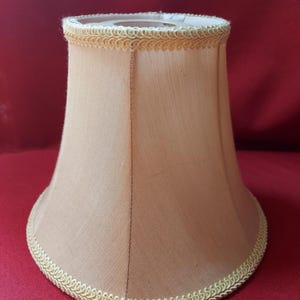 May include: A bell-shaped lamp shade in a light beige fabric with a smooth texture. The top and bottom edges are finished with a decorative, braided cream-colored trim. The shade features vertical seams and a circular opening at the top.