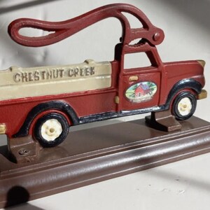 May include: A decorative cast iron truck in red, cream, and black, with the words "Chestnut Creek" on the side. The truck has a farm scene on the door and is mounted on a brown base. The truck is approximately 10 inches long.