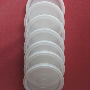 May include: A stack of eight white plastic lids, each with a slightly raised edge and a central indentation. The lids are arranged vertically against a red background, suggesting a kitchen or food storage context.