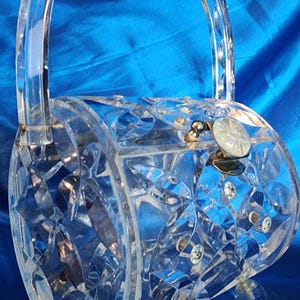 Pode incluir: Bolsa transparente em estilo vintage com alça curva. A bolsa é feita de material transparente facetado, com um fecho decorativo e pequenos pés de bola transparentes. O design da bolsa apresenta padrões em forma de diamante.