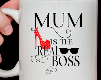 Può includere: Tazza in ceramica bianca con un design nero e rosso. La tazza dice "Mum is the real boss" con una scarpa con tacco alto rossa con pois bianchi e occhiali da sole neri.