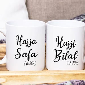 May include: Two white ceramic mugs with black calligraphy text. One mug reads "Hajja Safa Est. 2025" and the other reads "Hajji Bilal Est. 2025". The mugs are on a wooden tray.