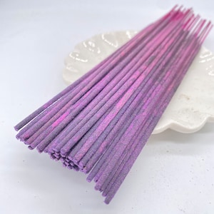 May include: A bundle of purple incense sticks with a glittery finish. The sticks are arranged on a white ceramic plate.