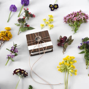 Könnte beinhalten: Ein Holznotizbuch mit dekorativem Knopf und Schnur, umgeben von verschiedenen bunten Blumen. Das Notizbuch hat ein weißes Etikett mit Text. Zu den Blumen gehören lila, gelbe und rosa Blüten, die eine lebendige, natürliche Ästhetik schaffen.