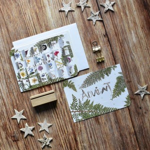 Könnte beinhalten: Ein Adventskalender mit kleinen Fenstern, die mit getrockneten Blumen gefüllt sind, auf einer Holzoberfläche. Das Wort "Advent" ist auf einer Karte mit Farnblatt-Akzenten geschrieben. Dekorative Sterne und goldene Büroklammern sind ebenfalls vorhanden.