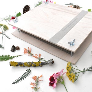Könnte beinhalten: Eine Holzbuchpresse mit einem grauen Stoffstreifen und dem aufgedruckten Text "meine große Blumencollage". Die Presse ist von verschiedenen Blumen, Blättern und Tannenzapfen umgeben.