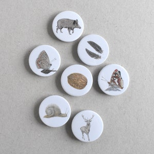 Könnte beinhalten: Sieben weiße, runde Buttons mit Illustrationen aus der Natur. Die Bilder zeigen ein Wildschwein, Federn, einen Schmetterling, eine Walnuss, einen weiteren Schmetterling, eine Schnecke und ein Reh. Die detaillierten Illustrationen sind in Graustufen gehalten.