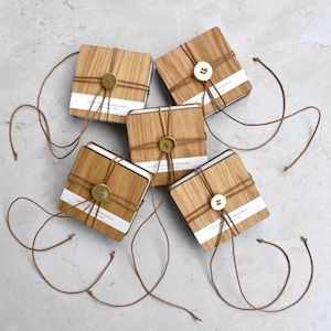 Könnte beinhalten: Fünf quadratische Untersetzer aus hellem Holz sind auf einer hellgrauen Oberfläche angeordnet. Jeder Untersetzer ist mit Bindfaden umwickelt und mit einem goldfarbenen Knopf befestigt. Auf jedem Untersetzer ist ein weißes Etikett mit Text zu sehen.