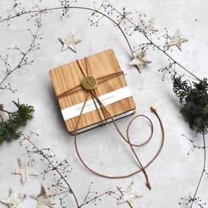 Könnte beinhalten: Eine quadratische Holzbox mit hellbraunem Finish, mit Bindfaden und einem großen Knopf gebunden. Ein weißes Etikett mit Text ist sichtbar. Die Box ist von dekorativen Sternen und Zweigen auf einer hellgrauen Oberfläche umgeben.