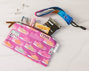 May include: A pink zippered pouch with a running shoe pattern. The pouch is open and contains a pink and yellow Emergen-C packet, a brown protein bar, a clear lip balm, and a blue and purple wrist strap.