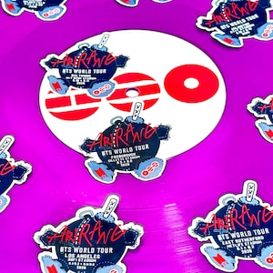 May include: A bright purple vinyl record featuring multiple die-cut stickers shaped like dark blue teddy bears with red text that reads Arirang BTS World Tour. The center label is white with three red horizontal bars and a red circle design.