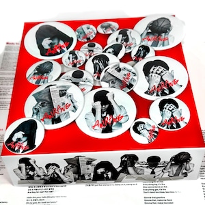 Puede incluir: Una colección de chapas redondas con imágenes fotográficas en blanco y negro de personas con los rostros ocultos, acentuadas por el texto rojo Arirang. Las chapas se muestran sobre una caja roja con imágenes a juego.