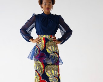 May include: A woman wearing a navy blue long-sleeved blouse with a high neck and a patterned skirt. The skirt is a dark blue base with red, yellow, and white abstract patterns. The skirt has a sheer overlay at the top.