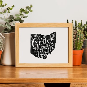 Op de afbeelding: Een rechthoekige lijst van licht hout met een witte print met een zwarte silhouet van de staat Ohio. Het silhouet bevat de witte handgeschreven tekst With God All things are Possible gecentreerd in de vorm van de staat.
