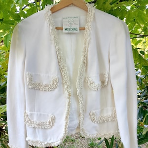 Peut inclure: Veste blanc cassé avec garniture et poches à volants. La veste est suspendue à un cintre en bois sur un fond de feuillage vert. L'étiquette indique "MOSCHINO".