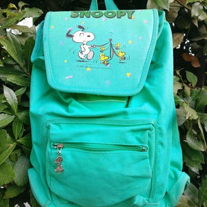 May include: A bright green backpack featuring a graphic of Snoopy and Woodstock on the top flap with the text SNOOPY printed above. The bag includes a front zippered pocket with a metal Snoopy character zipper pull and adjustable shoulder straps.