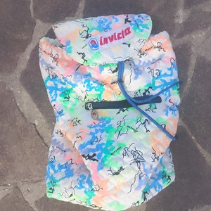 May include: A quilted Invicta backpack featuring a white base with a multi-colored abstract pattern in neon pink, blue, and green. The bag includes a top flap with the Invicta logo, a dark blue zippered front pocket, and blue drawstring cords.